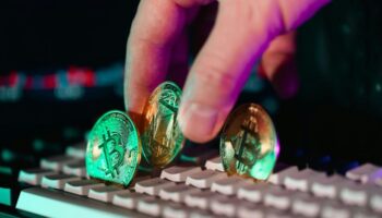 Mão segurando moedas físicas de Bitcoin e Ethereum sobre um teclado de computador.