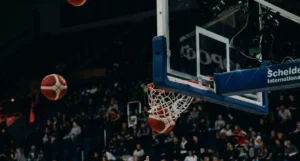 Bola de basquete caindo dentro da rede de uma cesta durante um jogo em um ginásio repleto de torcedores.